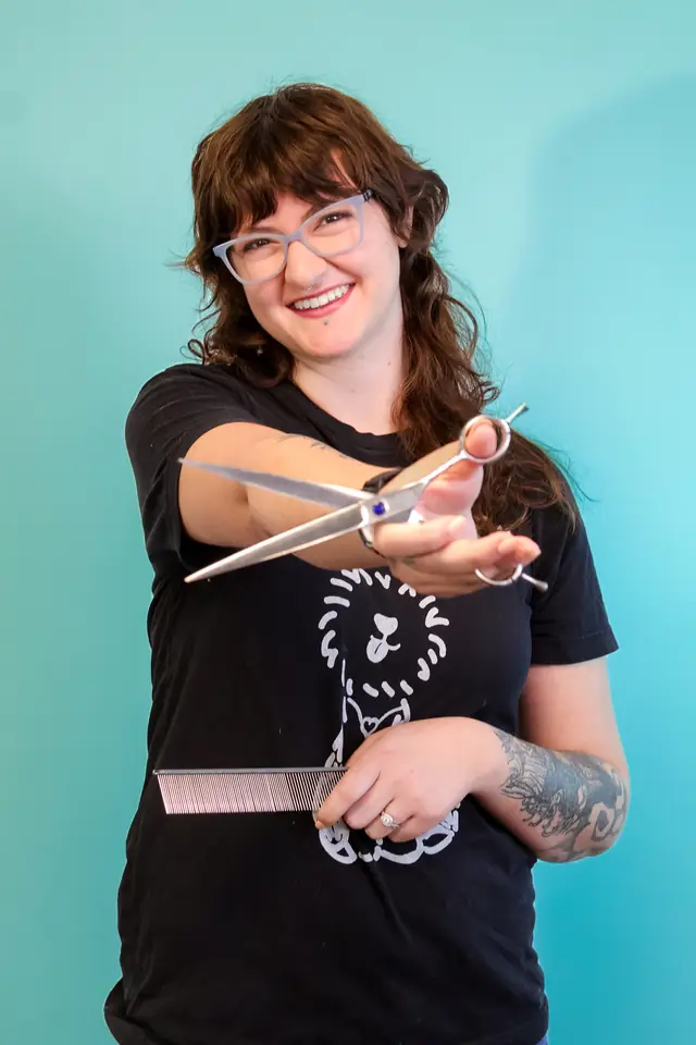 A groomer in a black Bark Me Beautiful tee holding grooming scissors and a comb