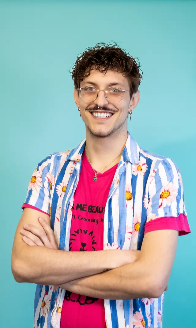 A team member in a patterned shirt with arms crossed, smiling, against the turquoise wall